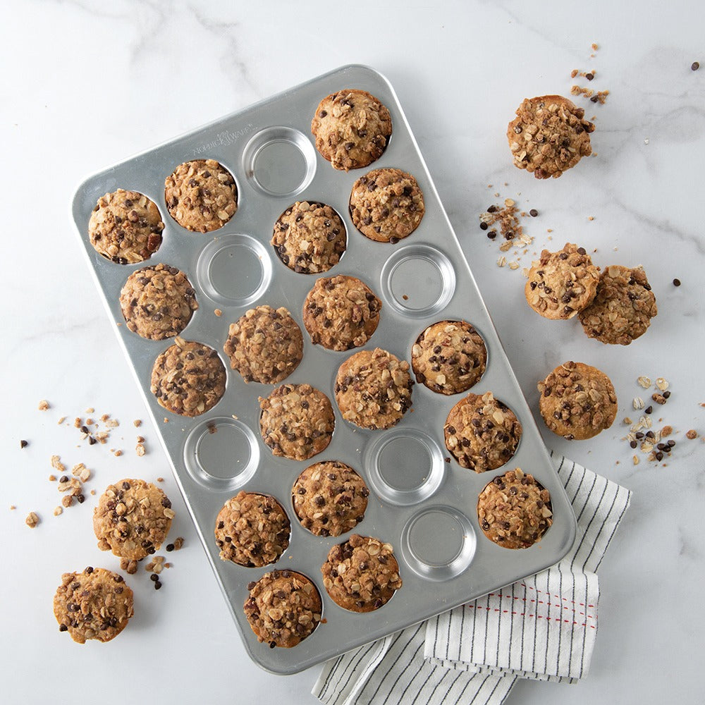 Oat chocolate-chip muffins in a silver 24-well muffin pan with scattered crumbs